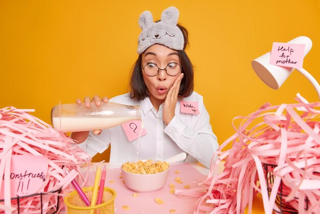 vrouw staart naar fles melk en cornflakes kan niet geloven dat haar ogen poseert op rommelig bureaublad werkt vanuit huis draagt blinddoek op voorhoofd geïsoleerd op geel