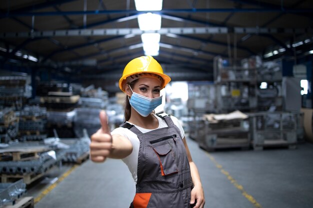 Vrouw staat in de fabriekshal en duimen opdagen terwijl ze een hygiënisch masker draagt als preventie tegen het coronavirus