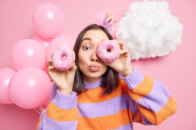 vrouw tegen ogen met smakelijke donuts houdt lippen gevouwen gekleed in casual trui viert verjaardag geniet van feestelijke gelegenheid geïsoleerd op roze