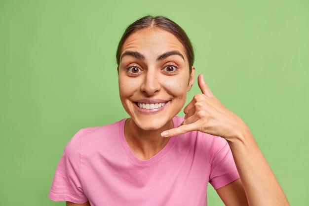 vrouw toont gebaar van mobiele telefoon bel me bord vraagt om telefoonnummer voelt zich tevreden draagt casual roze t-shirt op groen