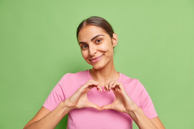 vrouw toont hart liefdesgebaar deelt haar gevoelens met jou glimlacht zacht gekleed in casual roze t-shirt op groen