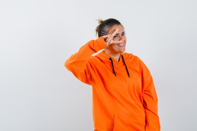 Vrouw toont V-teken op oog in oranje hoodie en kijkt vrolijk