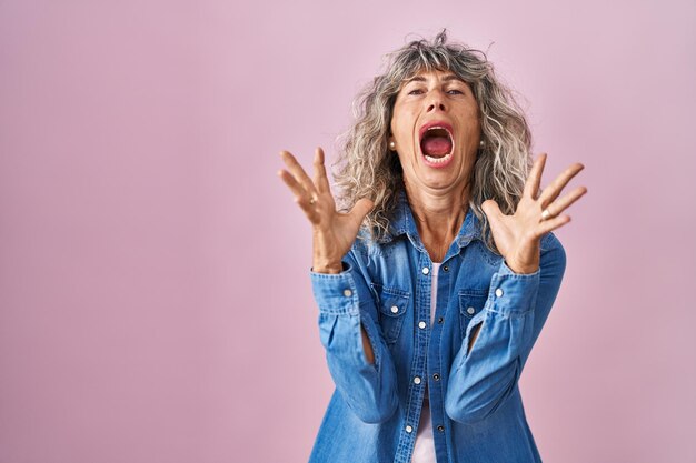 Vrouw van middelbare leeftijd die over een roze achtergrond staat, gek en gek schreeuwt en schreeuwt met agressieve uitdrukking en opgeheven armen. frustratie concept.