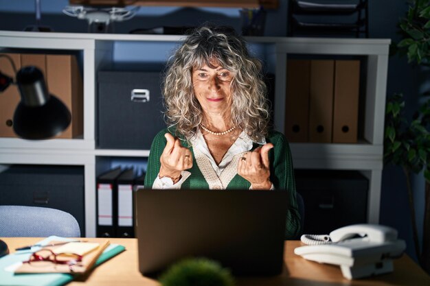 Vrouw van middelbare leeftijd die 's nachts werkt met behulp van een computerlaptop die geld gebaart met de handen, om salarisbetaling vraagt, miljonair