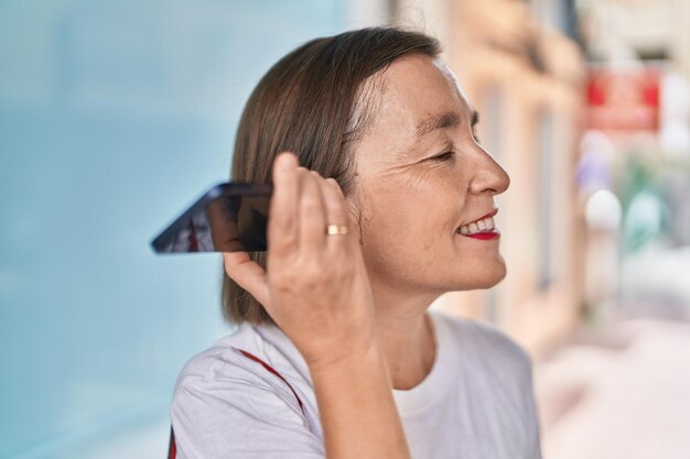 Vrouw van middelbare leeftijd glimlachend zelfverzekerd luisterend audiobericht door de smartphone op straat