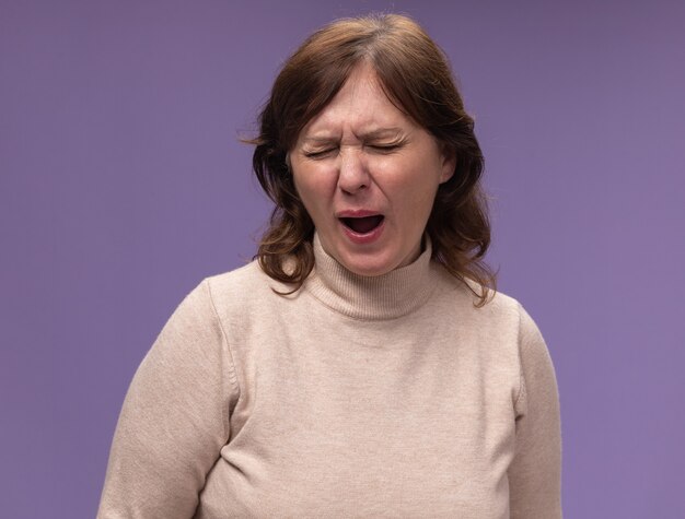 Vrouw van middelbare leeftijd in beige coltrui op zoek moe en verveeld geeuwen staande over paarse muur