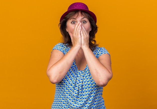 Vrouw van middelbare leeftijd in feestmuts die geschokt is over de mond met handen die een verjaardagsfeestje vieren dat over de oranje muur staat