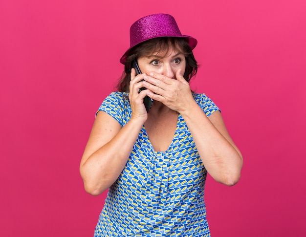 Vrouw van middelbare leeftijd in feestmuts die verbaasd kijkt terwijl ze de mond bedekt met de hand terwijl ze op een mobiele telefoon praat