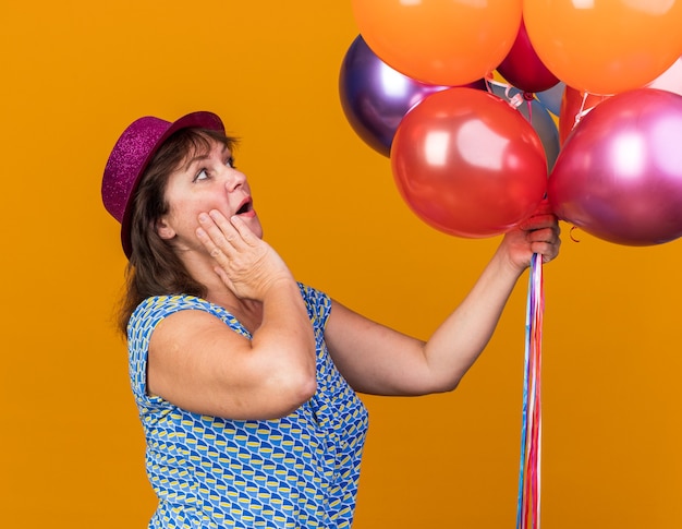 Vrouw van middelbare leeftijd in feestmuts met kleurrijke ballonnen die naar hen kijkt verbaasd en verrast om een verjaardagsfeestje te vieren dat over de oranje muur staat