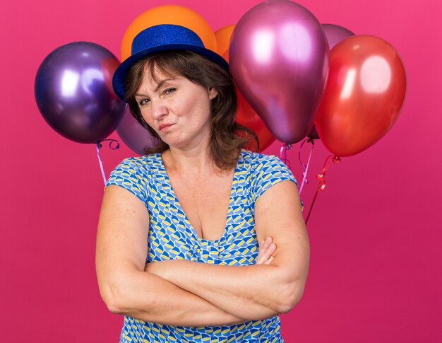 Vrouw van middelbare leeftijd in feestmuts met kleurrijke ballonnen die ontevreden is met gekruiste armen om verjaardagsfeestje te vieren dat over roze muur staat