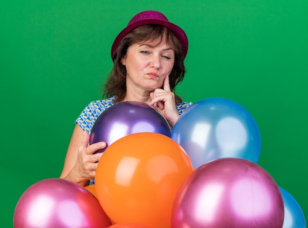 Vrouw van middelbare leeftijd in feestmuts met kleurrijke ballonnen met een sceptische uitdrukking die een verjaardagsfeestje viert dat over de groene muur staat