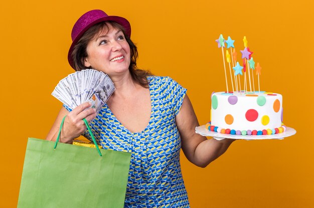 Vrouw van middelbare leeftijd in feestmuts met papieren zak met geschenken met verjaardagstaart en contant geld blij en tevreden glimlachend vrolijk verjaardagsfeestje vieren staande over oranje muur