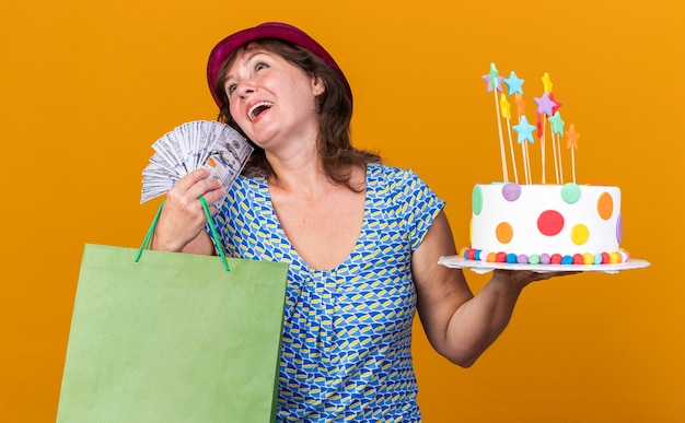 Vrouw van middelbare leeftijd in feestmuts met papieren zak met geschenken met verjaardagstaart en contant geld blij en tevreden glimlachend vrolijk verjaardagsfeestje vieren staande over oranje muur