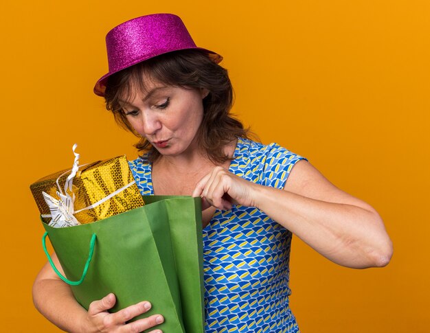 Vrouw van middelbare leeftijd in feestmuts met papieren zak met verjaardagscadeaus die er geïntrigeerd uitziet om een verjaardagsfeestje te vieren dat over de oranje muur staat