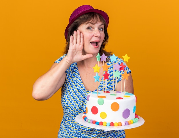 Vrouw van middelbare leeftijd in feestmuts met verjaardagstaart met hand over oor die roddels probeert te horen
