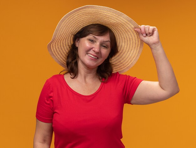 Vrouw van middelbare leeftijd in rode t-shirt en zomerhoed blij en positief glimlachend vrolijk staande over oranje muur