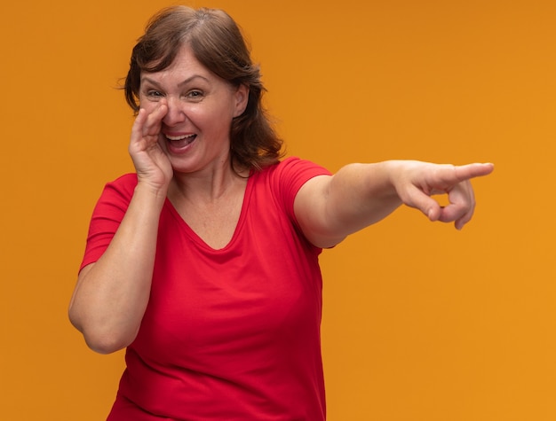 Vrouw van middelbare leeftijd in rood t-shirt glimlachend vertellend een geheim met hand dichtbij mond die met wijsvinger richt op iets dat zich over oranje muur bevindt