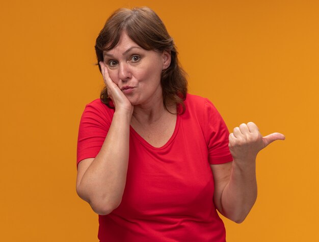 Vrouw van middelbare leeftijd in rood verbaasd t-shirt wijzend met duim naar de kant die zich over oranje muur bevindt