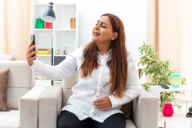 Vrouw van middelbare leeftijd in wit overhemd en zwarte broek selfie met smartphone gelukkig en zelfverzekerd zittend op de stoel in lichte woonkamer