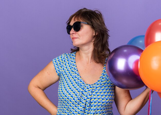 Vrouw van middelbare leeftijd met een bril die een stel kleurrijke ballonnen vasthoudt en opzij kijkt met een serieus gezicht dat een verjaardagsfeestje viert dat over een paarse muur staat
