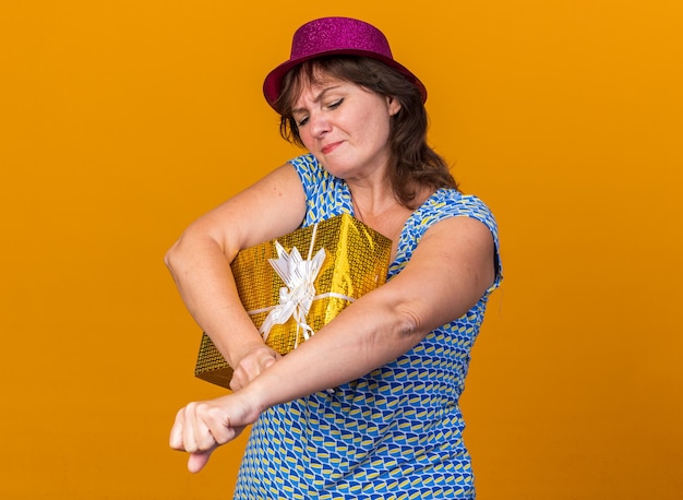 Vrouw van middelbare leeftijd met een feesthoed die een cadeau vasthoudt en er verward en ontevreden uitziet om een verjaardagsfeestje te vieren dat over de oranje muur staat