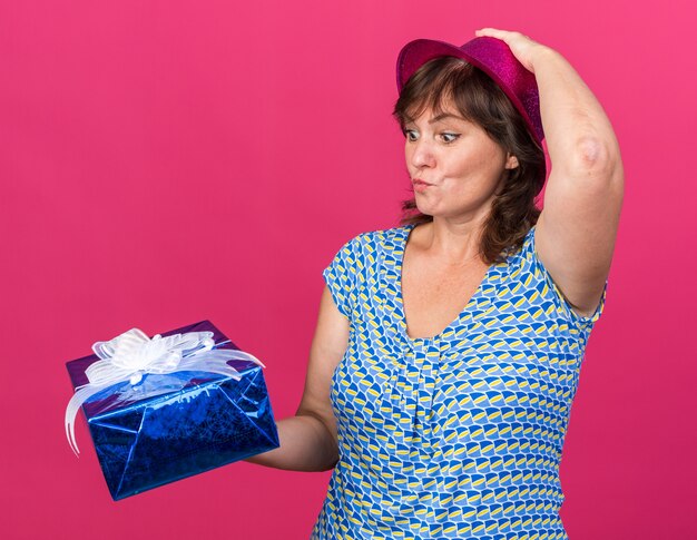 Vrouw van middelbare leeftijd met een feestmuts die een cadeautje vasthoudt en ernaar kijkt verward met haar hand op haar hoofd die een verjaardagsfeestje viert dat over een roze muur staat