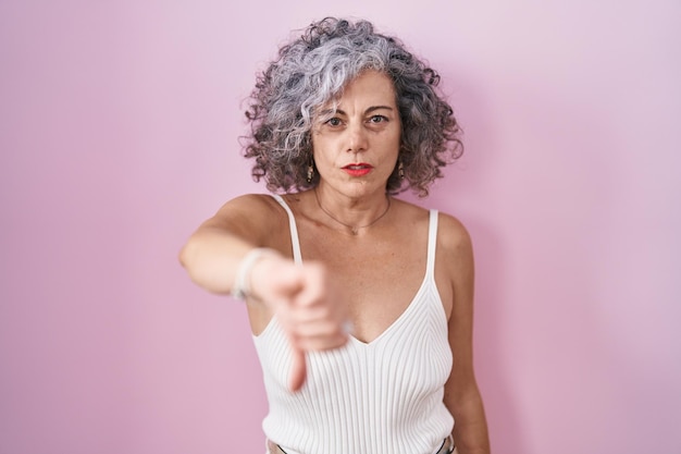 Vrouw van middelbare leeftijd met grijs haar dat over een roze achtergrond staat en er ongelukkig en boos uitziet en afwijzing en negatief toont met een duim omlaag gebaar. slechte uitdrukking.