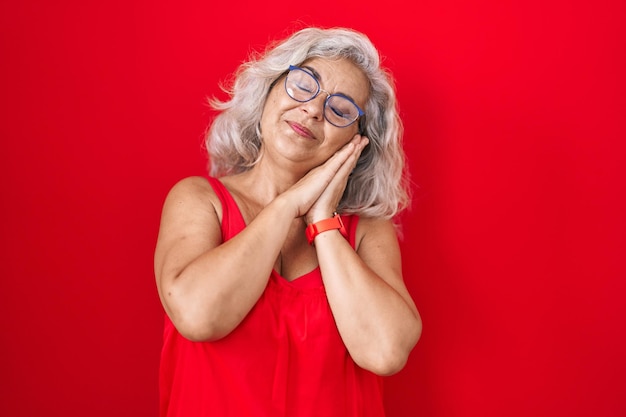 Gratis foto vrouw van middelbare leeftijd met grijs haar die over een rode achtergrond staat, moe droomt en poseert met de handen in elkaar terwijl ze glimlacht met gesloten ogen