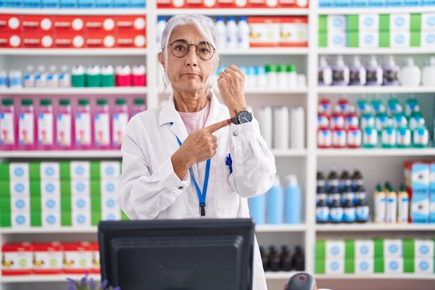 Vrouw van middelbare leeftijd met tatoeages werken bij apotheek drogisterij in haast wijzend om naar de tijd te kijken, ongeduld, kijkend naar de camera met een ontspannen uitdrukking