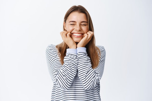 Vrouw verheugt zich over goed nieuws, houdt handen op het gezicht en lacht met gesloten ogen, juicht en viert, hoort iets positiefs, staat over een witte muur