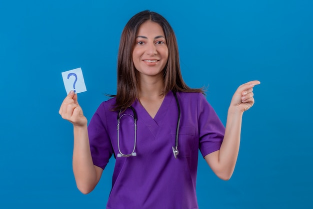 Vrouw verpleegster in medisch uniform en met stethoscoop met herinneringspapier met vraagteken erg blij wijzend met hand en vinger naar de zijkant staande op blauw