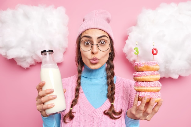 vrouw viert 30e verjaardag houdt geglazuurde heerlijke donuts met verse melk houdt lippen gevouwen draagt vrijetijdskleding geïsoleerd op roze