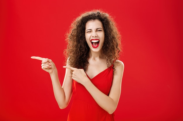 Vrouw voelt zich geweldig op een geweldig feest van een vriend die naar de ingang wijst. Stijlvolle zorgeloze vrouw met krullend haar in rode avondjurk die plezier heeft in de buurt van het zwembad dat vrolijk glimlacht en naar links wijst.