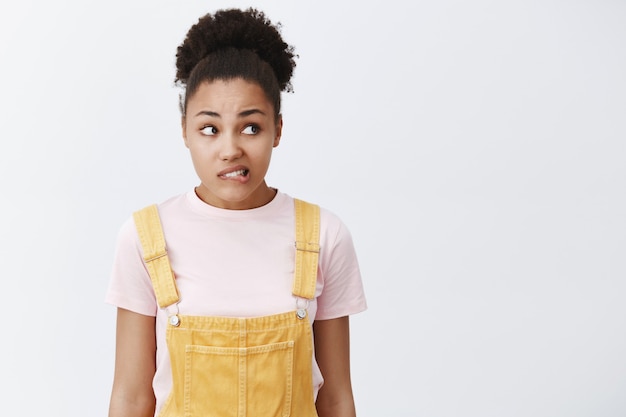 Vrouw voelt zich schuldig, wil sorry zeggen. portret van nerveus en bezorgd schattig afrikaans amerikaans meisje in gele overall, lip bijten en angstig naar rechts staren, staande over grijze muur