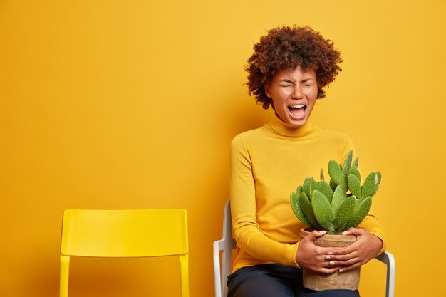 vrouw voelt zich wanhopig houdt pot met cactus houdingen op stoel op geel