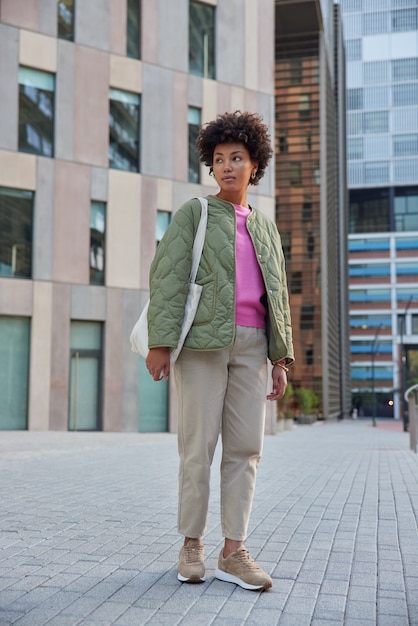 Vrouw wandelt in de stad tegen stadsgebouwen poseert in het centrum kijkt weg denkt aan iets heeft een excursie in het centrum