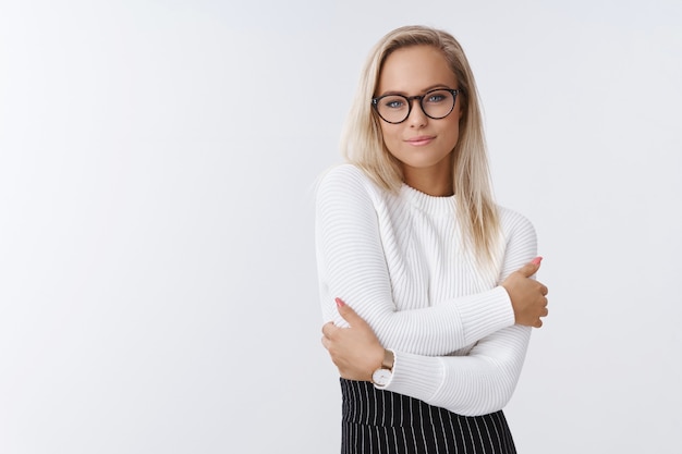 Vrouw weet hoe elegant en warm aankleden in de winter op kantoor op een witte achtergrond in trendy trui en bril knuffelt, zichzelf knuffelt en zich gezellig en comfortabel voelt glimlachend zacht.