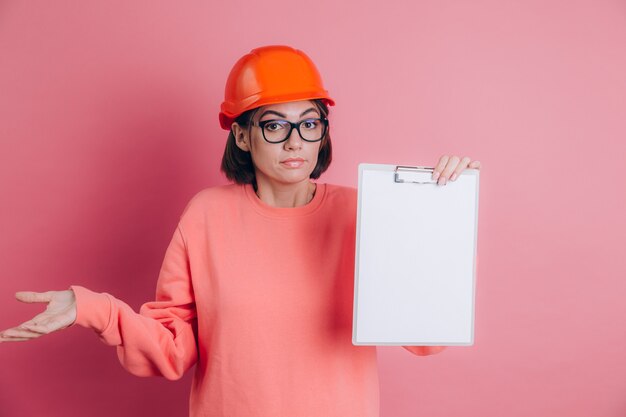 Vrouw werknemer bouwer houdt witte bord leeg tegen roze achtergrond. Helm bouwen. Vrouw werpt haar handen op en haalt haar schouders op, zonder enig idee