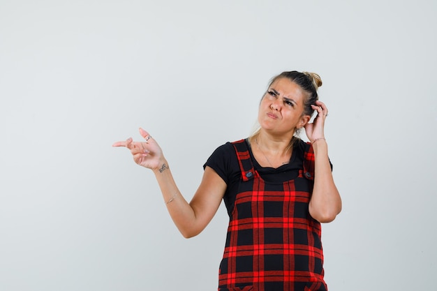 Vrouw wijst opzij, hoofd krabben in overgooier jurk en op zoek aarzelend, vooraanzicht.