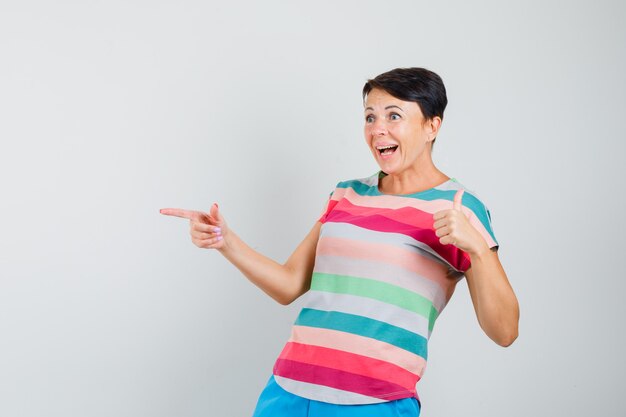 Vrouw wijst opzij, toont duim in gestreept t-shirt, broek en kijkt verbaasd, vooraanzicht.