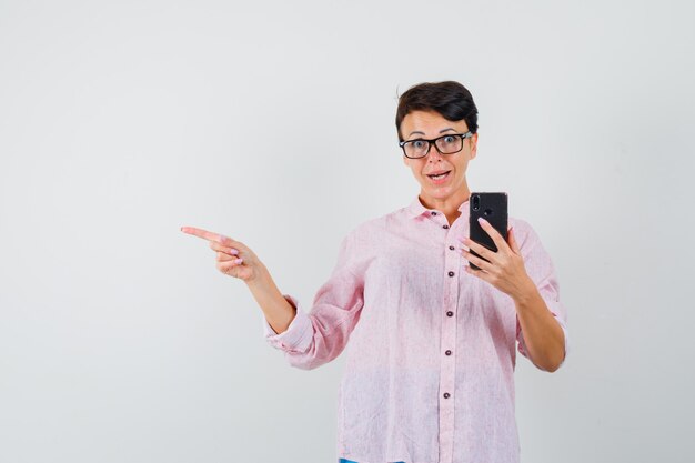 Vrouw wijst weg terwijl ze mobiele telefoon in roze shirt vasthoudt en opgewonden kijkt, vooraanzicht.