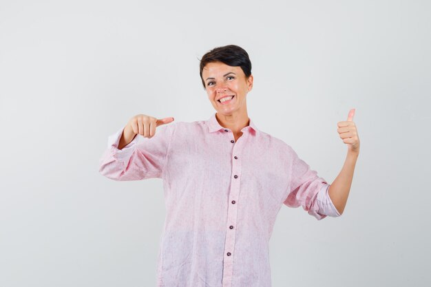 Vrouw wijzend op haar duim omhoog in roze shirt en op zoek vrolijk. vooraanzicht.