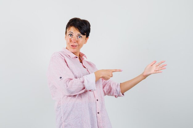 Vrouw wijzend op haar gespreide handpalm in roze shirt en kijkt verward, vooraanzicht.
