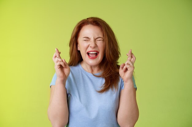 Vrouw wil winnen erg enthousiast gelukkig roodharige vrouw van middelbare leeftijd smeken smeken god laat droom uitkomen kruis vingers veel geluk wensen gesloten ogen open mond opwinding groene muur