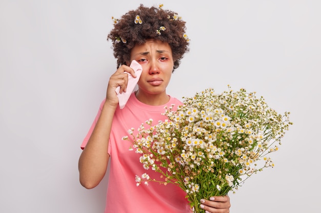 Gratis foto vrouw wrijft rode tranende ogen met tissues houdt een boeket kamillebloemen vast die allergisch zijn voor stuifmeel geïsoleerd over wit