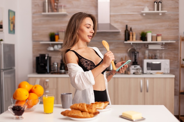 Vrouw zingen tijdens het ontbijt in de keuken van het huis, het dragen van sexy lingerie. Verleidelijke vrouw met tatoeages met smartphone die 's ochtends tijdelijk ondergoed draagt.