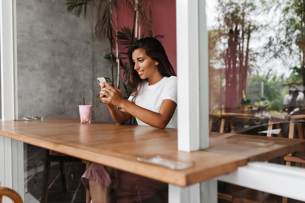 Vrouw zit aan tafel in een gezellig café en chats in smartphone Portret van gebruind meisje in wit Tshirt