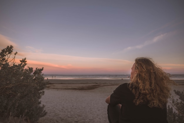 Gratis foto vrouw zitten in de buurt van de kust van de zee en kijken naar de zonsondergang