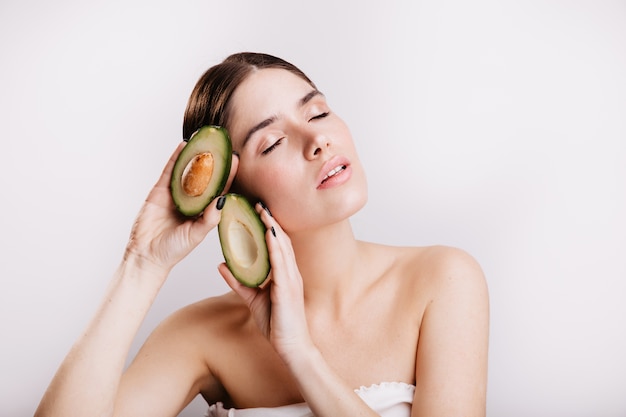 Vrouwelijk model met heldere huid geïnspireerd poseren op geïsoleerde muur met voedzame, gezonde avocado's.