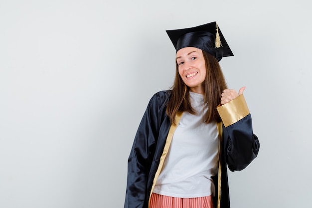 Vrouwelijke afgestudeerde duim opdagen in academische kleding en op zoek gelukkig. vooraanzicht.